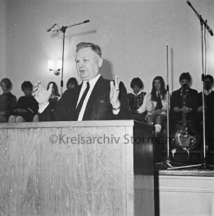Theodor-Mommsen-Schule: Aula: Theodor-Mommsen-Fest: vorn Rednerpult mit Redner: dahinter auf der Bühne sitzende Schülergruppe und Mikrofone: 7. Dezember 1968