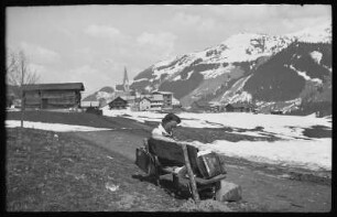 Lesende Frau (Edit Kammer?) auf einer Bank, im Hintergrund Mittelberg mit der Kirche St. Jodok im Kleinwalsertal