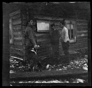 3 Offiziere mit Hund vor einer Schrifttafel am Eck eines Holzhauses, Schneereste
