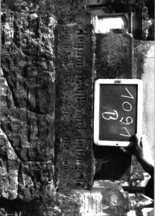 Grabstein 1091 (Index: 100104145) Material: Sandstein Zustand: teilweise angewittert Maße HxBxT: 178x53x20 cm Beschreibung: Stele auf getrepptem Sockel, Aufsatz mit Dreiecksabschluss und dahintergestellten spitzbogigen Giebel- sowie Eckakroterien, Giebelakroter mit Mohnkapseln, Eckakroterien mit Voluten, unterster Sockel felsartig gearbeitet, Stele wirkt unproportioniert;Mittelteil ist zu niedrig im Verhältnis zum Sockel Symbolik: Mohnkapseln, Voluten: Grabstein 1091B