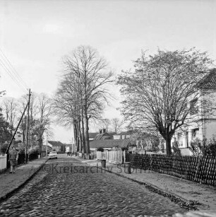 Hoisbütteler Dorfstraße: Kopfsteinpflaster: rechts auf Gehweg Mülltonnen: links Frau: dahinter Strommast: rechts und links Häuser mit Vorgärten, Zäunen und Bäumen