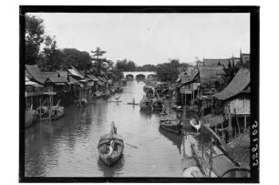 Fluss in Phetchaburi : Phetchaburi. Blick auf den von Häusern gesäumten Fluss mit Booten gegen eine Brücke, 1909