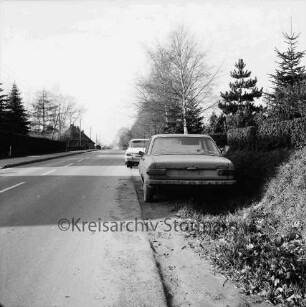 Wolkenweher Dorfstraße: vorn Schrottauto am Straßenrand: dahinter parkender Pkw: rechts und links Hecken und Bäume: hinten links Häuser