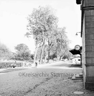 Bahnhofsgebäude: Straßenfassade: Uhr, Nebengebäude von Güterbahnhof: Bahnhofstraße mit Kopfsteinpflaster, Fahrradfahrer, Straßenbäume: an westlicher Straßenseite unbebautes Grundstück, Arbeiter, Hecke, Ruhebank