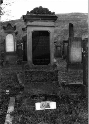 Grabstein 86 (Index: 99798547) Material: Sandstein, Schriftplatte Granit Zustand: gut, Schriftplatte gesprungen Maße HxBxT: 176x46x25 cm Beschreibung: Stele mit Volutenaufsatz, Schriftplatte mit geradem Aufsatz, Sockel: Grabstein 86