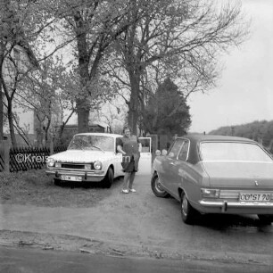 Meisenweg: Charlotte Mallek zwischen zwei parkenden Pkws: links Ecke eines Wohnblocks und Schuppen hinter Zaun und Bäumen: hinten rechts Mann und Bäume