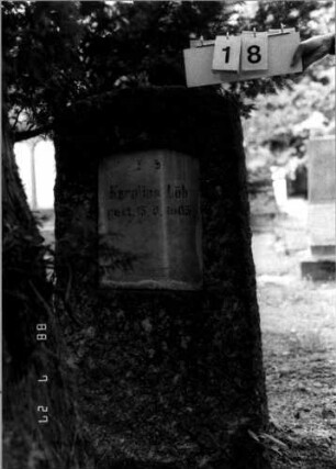 Grabstein 18 (Index: 100110205) Material: Granit mit weißer Marmorplatte Zustand: leicht verwittert Maße HxBxT: 124x60x32 cm Beschreibung: felsenartige Stele, Schriftplatte mit Segmentbogen: Grabstein 18