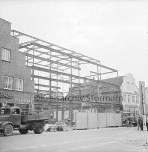 Firma Carl Hoffmann: Hindenburgstraße: Elektrohandel: Neubau: Aufbau des Stahlgerüsts: vorn Bauzaun, zwei Lkws, Bretterstapel und Fußgänger: rechts und links Wohn- und Geschäftshäuser mit Haushaltswarengeschäft Otto Möller und Elektrogeschäft Carl Hoffmann