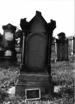 Grabstein 98 (Index: 99937117) Material: Sandstein Zustand: Sockel beschädigt Maße HxBxT: 158x52x30 cm Beschreibung: Stele mit Halbsäulen, getreppter Sockel, giebelartiger Abschluß mit Verdachung und Eckakroteren, Schriftfeld eingetieft mit Rundbogenabschluß, rechteckiges Schriftfeld im Sockel Symbolik: Ranken in den Akroteren: Grabstein 98