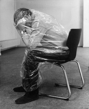 Einsamkeit. Ein Mann sitzt in Plastefolie eingepackt und abgeschirmt auf einem Stuhl.