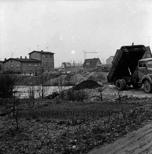 Baustelle mit Baugrube: vorn Büsche: dahinter Erdhaufen: rechts Lkw beim Abladen von Erde: hinten Ein- und Mehrfamilienhäuser: dahinter Gerüste und Baukran