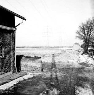 Feldmark: rechts Bäume und Erdhügel: links Gebäude: hinten Felder: darauf Strommasten der Überlandleitung: Winter