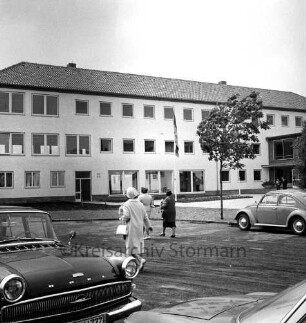 Ahrensburg: Hermann-Löns-Straße: Erweiterungsbau: Einweihung: Außenansicht: Gäste auf Straße und Vorplatz: Fahnenmast: hinten rechts Verbindungstrakt zum Altbau: im Vordergrund Pkw, 26. Juni 1962