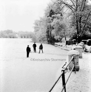 Mühlenteich mit Eis- und Schneedecke: darauf drei Schlittschuhläufer: links Geländer: dahinter Bäume: Winter
