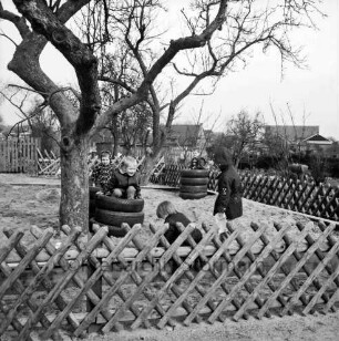 Kindergarten in ehemaligem Volksschulgebäude: Schulstraße: Hof mit Spielgerät aus aufeinandergestapelten Autoreifen: darin spielende Kinder: vorn Baum und Jägerzaun: hinten Wohnhäuser