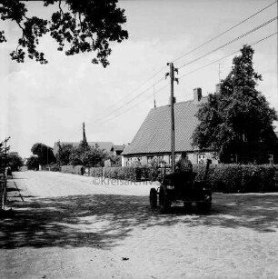 Hauptstraße mit Kopfsteinpflaster: rechts Trecker: dahinter Bauernhäuser mit Vorgärten und Hecken: Strommasten