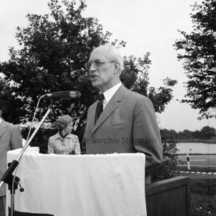 Haarmann, Wennemar: am Rednerpult vor Mikrofon: links zwei Personen: hinten rechts Straße: dahinter See: hinten Bäume