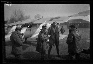 Nicht identifizierbare Ortschaft in Nordfrankreich oder Belgien: 3 Piloten in Fliegerjacken im Gespräch, im Hintergrund ein Doppeldecker mit Tatzenkreuz auf den Tragflächen und am Rumpf und ein Hangar ("Jagdstaffel Czermak")