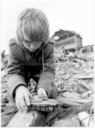 Junge kniet auf einem Schutthaufen und spielt mit seinem Miniaturpanzer (Altersgruppe 14-17)