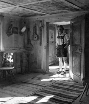 Bauernstube. Interieur mit geschnitztem Stuhl und Saiteninstrumenten an der Wand. In der göffneten Tür ein junger Mann in Lederhosen (vielleicht im Allgäu)