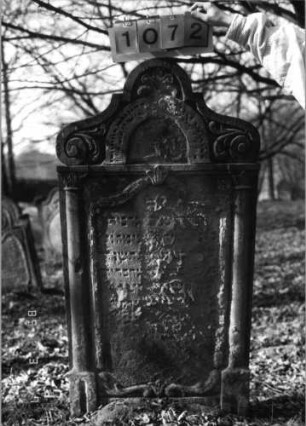 Grabstein 1072 (Index: 99753331) Material: Sandstein Zustand: abgeblättert/fast völl. Textverlust Maße HxBxT: 138x72x10 cm Beschreibung: ädikulaart. St. m. Halbsäulen, in Voluten auslaufender Rundbogenaufsatz Symbolik: Ranken, Girlande, Voluten: Grabstein 1072
