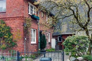 Backsteinhaus mit umlaufendem Gesims: Giebelseite [angeschnitten], Traufseite mit Sprossenfenstern, Rankgittern: rechts Querbau, davor Wasserpumpe: Plattenweg, vorne Gartenzaun, Gartenpforte aus Schmiedeeisen
