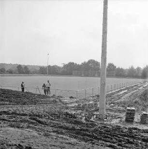 Lottbek: Sporteinrichtungen: Volksdorfer Weg: Sportplatz vor Fertigstellung: vorn rechts Stapel mit Gehwegplatten: daneben Mast für Flutlicht: hinten links an Geländer drei Arbeiter mit Schaufeln: hinten Trennnetz und Bäume