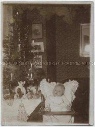 Baby im Kinderstuhl vor Weihnachtsbaum und Gabentisch