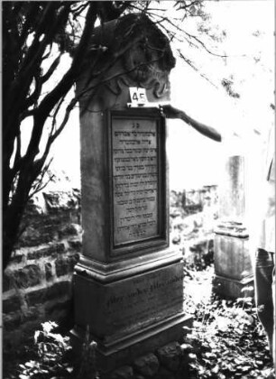 Grabstein 45 (Index: 99804406) Material: Sandstein Zustand: Sockel leicht abgeblättert Maße HxBxT: 214x59x26 cm Beschreibung: Stele, getreppter profilierter Sockel, Rundbogenabschluß mit Verdachung, Schriftfeld rechteckig Symbolik: Kranz mit Schleife: Grabstein 45