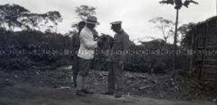 Der Leiter der Station Molundu und ein Angehöriger der Schutztruppe in Uniform mit kleinen Affen auf dem Arm, im Hintergrund ein afrikanischer Polizeisoldat