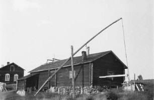 Blockhaus und Ziehbrunnen : Blockhaus in einer Siedlung. Davor ein Ziehbrunnen.