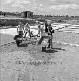 Freibad: Reeshoop: in Bau: Arbeiter mit Schubkarren beim Aufbringen der Fundamentierung für Sohle von Nichtschwimmerbecken: hinten Holzschuppen, Leiter, Zuschauer, Bauzaun