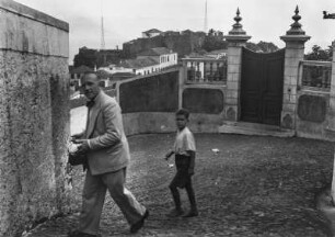 Straßenszene : Ein Mann und ein Junge in einer Straße. Hinter ihnen eine Mauer mit Tor, im Hintergrund weitere Gebäude und zwei Antennen.