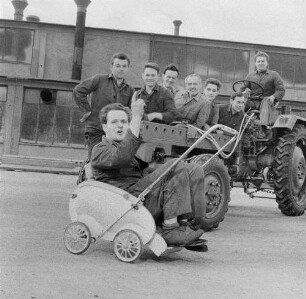 Schönebeck. VEB Traktorenwerk Schönebeck. Produktionsbereich Geräteträger RS 09. Gruppe Kraftfahrzeugschlosser an und auf einem Fahrzeug auf dem Werkgelände, davor ein Schlosser in einem Kinderwagen sitzend