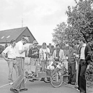 Vatertagstour: Männer mit Biergläsern bei zweirädrigem Karren mit Bierfass, Lautsprecher, Korb: hinten Haus