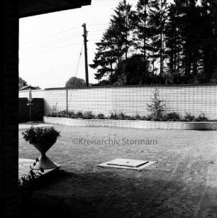 Gutsgärtnereigelände: Vorplatz vor Bungalow: rechts Blumenschale: hinten Mauer: davor Blumerabatten: hinten links Sonnenschirm: hinter der Mauer Hausdach, Strommast und Bäume