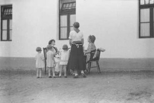 Frauen und Kinder : Um einen Tisch drei Frauen und drei kleine Mädchen; hinter ihnen ein großes Haus.