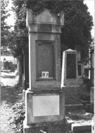 Grabstein 87 (Index: 100114219) Material: Kalkstein mit weißer Marmorplatte im Sockel Zustand: obere Schriftplatte fehlt Maße HxBxT: 210x66x37 cm Beschreibung: Stele, getreppter Sockel, Giebel mit Eckakroteren, eingetieftes Schriftfeld und Schriftplatte im Sockel rechteckig: Grabstein 87