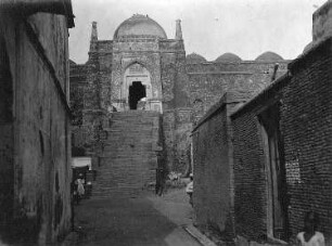 Delhi, Indien. Jama Masjid (Masjid-i Jahan Numa, "Freitagsmoschee") (1644-1658; Shah Jahan). Blick durch eine Gasse mit Wohnhäusern auf einen Zugang zur Freitagsmoschee in massiver Festungsmauer mit Kuppel und Treppenstufen