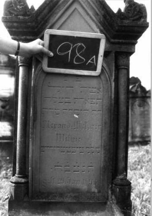 Grabstein 98 (Index: 99937117) Material: Sandstein Zustand: Sockel beschädigt Maße HxBxT: 158x52x30 cm Beschreibung: Stele mit Halbsäulen, getreppter Sockel, giebelartiger Abschluß mit Verdachung und Eckakroteren, Schriftfeld eingetieft mit Rundbogenabschluß, rechteckiges Schriftfeld im Sockel Symbolik: Ranken in den Akroteren: Grabstein 98A
