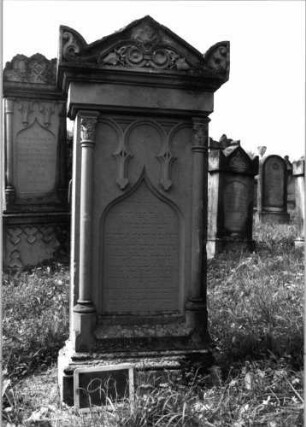 Grabstein 99 (Index: 99937115) Material: Sandstein Zustand: gut Maße HxBxT: 170x61x30 cm Beschreibung: ädikulaartige Stele mit Halbsäulen, profilierter Sockel, Giebel mit stilisierten Eckakroteren, Schriftfeld eingetieft mit Rundbogenabschluß Symbolik: Maßwerk mit herabhängenden Herzen, Voluten und Efeuranken im Giebel: Grabstein 99