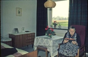 Kreisaltenheim [Reinfeld]: am Schwarzen Teich [später Kaliskaweg]: Innenaufnahme: Bewohner-Zimmer: Frau in Sessel beim Lesen von Buch