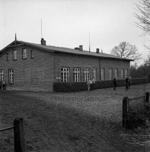 Volksschule: davor Hecke: links Schulkinder beim Spiel auf Schulhof: vorn Zaun und Eingang