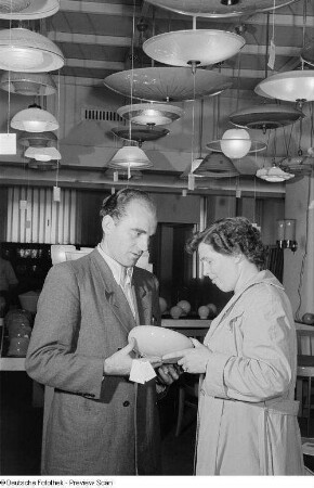 Ein Mann und eine Frau begutachten einen gläsernen Lampenschirm, im Hintergrund eine Vielzahl ausgestellter Deckenlampen der Leipziger Herbstmesse 1954