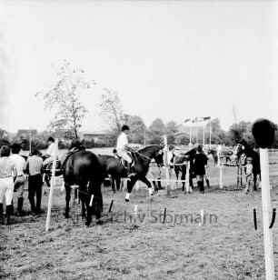 Sportveranstaltung: 2. Pferdeleistungsschau: Veranstalter Reitverein Ahrensburg/Hamburg-Ost: Reiter mit Pferden: Zuschauer an Zaun: im Vordergrund Absperrband, Reiterkappe auf Zaunpfahl: im Hintergrund Fahnenmasten mit Fahnen, Häuser