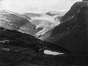 Gletscher : Blick auf die Gletscherzunge des Svartisen Gletschers. Auf einer Wiese im Vordergrund stehen zwei Wanderer