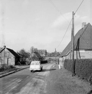 Dorfstraße mit Pkw: rechts Hecke und Strommast: dahinter Bauernhaus: hinten weitere Häuser und Bäume