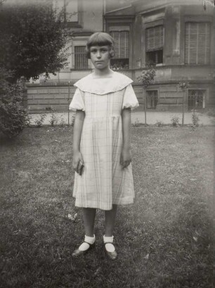 Mädchen im Sommerkleid auf einem Rasenstück vor Wohnhaus stehend