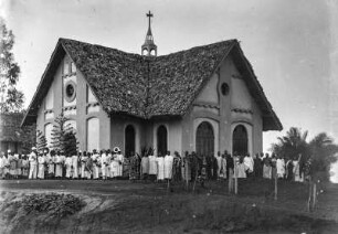 Kirchenweihe : Die Kirche der Mission in Mtai am Tag der Einweihung. Vor dem Gebäude stehen zahlreiche Personen, darunter auch links eine Blaskapelle mit Musikinstrumenten.