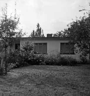 Behelfsheim: davor Rasenfläche, Blumenrabatten und Bäume